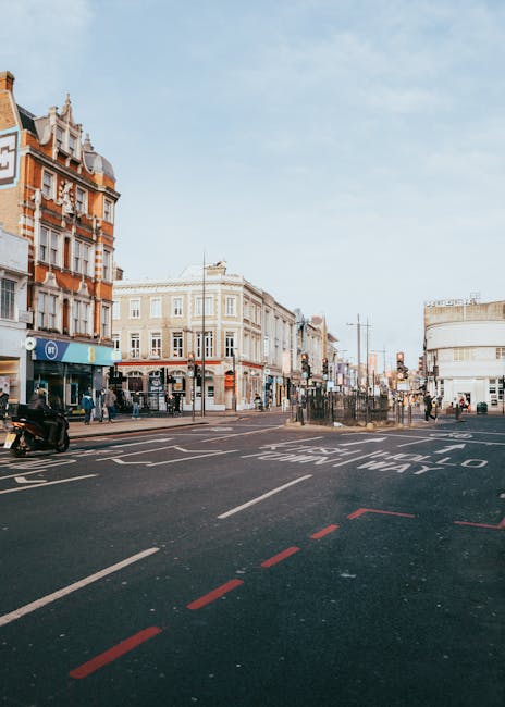 A wide view of Kilburn High Road with multi-storey and low-rise buildings featuring historic architectural details, including ornate facades and large windows, situated along the street on a clear day. The scene shows a busy urban environment with pedestrians walking on the sidewalks, some near shop entrances, and a few vehicles, including a parked motorcycle close to the curb. The road has designated parking and loading zones marked with red dashed lines and safety markings, indicating spaces used for moving and furniture transport during home relocation. Street furniture such as lampposts, traffic lights, and bus stops are visible, with an overall atmosphere of active city life. The lighting suggests natural daylight, possibly in the morning or early afternoon. This setting could be relevant to house removals or packing and moving services provided by Brent Removals, especially when considering logistics and loading processes in urban areas like Brent.