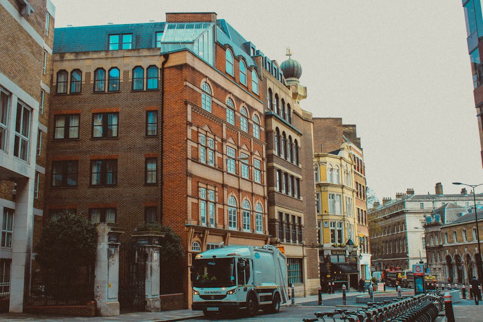 A street scene during daytime showing a white moving truck with a small orange light on top parked on the curb in front of a residential building with red brick and beige facade, large arched windows, and a teal-colored rooftop. The truck is positioned near the entrance of an apartment, which is partly obscured by a black metal gate. Several cardboard boxes, some wrapped in plastic or fabric packing materials, are visible inside the truck and on the pavement nearby, indicating a furniture transport or home relocation process. The sidewalk is lined with metal bollards, and there are other historic and modern buildings in the background. The scene captures the loading process typical of professional removals carried out by companies like Brent Removals, with a focus on the logistics of furniture and box transport on an urban street.