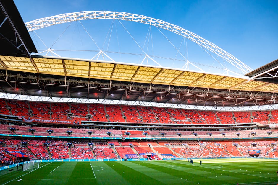 The exterior of a large football stadium during daytime, with a clear blue sky overhead. The stadium features a prominent white arch structure spanning across the roof, which is partially covered with a yellowish canopy. Inside, the seating area is visible with rows of red, pink, and purple seats, some of which are occupied by spectators. The lush green football pitch is in the foreground, with a white goalpost and net situated at one end. Several individuals are seen walking or standing on the pitch, possibly staff involved in a home relocation or event setup. Equipment such as trolleys and moving blankets may be present near the entrance. The scene suggests preparations for a match or event, with activity related to loading or setting up furniture, boxes, or other equipment typical of a house or venue move, conveying the process of furniture transport and packing in a stadium setting, aligned with Brent Removals' services.