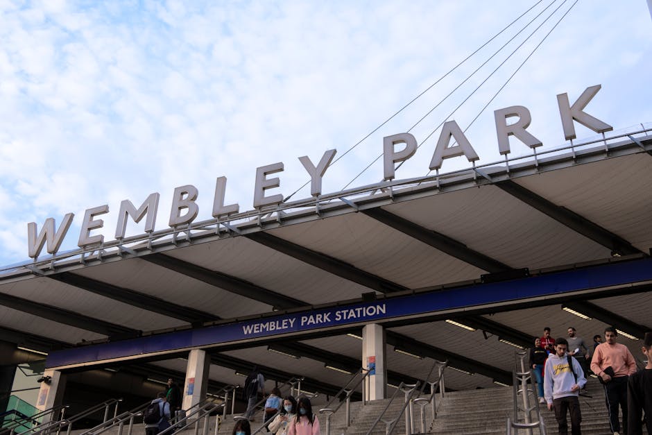 A wide-angle view of the exterior entrance to Wembley Park Station during daytime, featuring large, white, three-dimensional letters spelling 'WEMBLEY PARK' mounted on the roof overhang. Beneath the sign, there is a blue sign displaying 'WEMBLEY PARK STATION' in white text, attached to the station's structure. The image shows multiple people exiting or entering the station via concrete stairs, some wearing face masks, indicating a busy scene typical of home relocation or moving logistics. The station's canopy is supported by black metal beams, and overhead electrical wires run above the station entrance. The sky is partly cloudy with patches of blue visible, and the environment suggests an active transportation hub suitable for coordinating furniture transport and moving services, as facilitated by companies like Brent Removals.