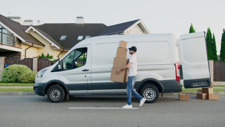 A man stands inside the open rear of a large moving van, which contains numerous stacked cardboard boxes of various sizes, some wrapped in plastic or packing materials. The man, dressed casually in a 
